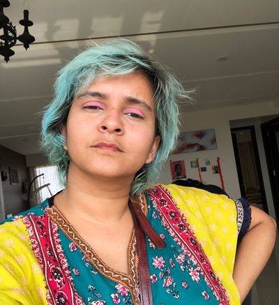Rajvi Desai, a brown non-binary person, strares at the camera with slightly closed eyes. She has teal hair with black roots, pink eyeshadow and a brightly patterned shirt. One arm rests akimbo while the other is out of frame.