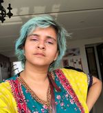 Rajvi Desai, a brown non-binary person, strares at the camera with slightly closed eyes. She has teal hair with black roots, pink eyeshadow and a brightly patterned shirt. One arm rests akimbo while the other is out of frame.