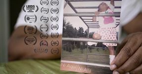 A closeup of a printed photograph of filmmaker Ley Comas as a young Black child joyously climbing monkey ladders in a backyard. The picture is being held by Ley Comas in the present, with their fingers and arm in the frame as well as part of their white t shirt. Numerous festival accolades are presented on the left side of the frame beside the photograph.