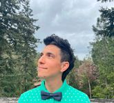 Kalil Cohen looks off into the distance against a forrested landscape. Kalil has short wavy dark hair with a few white strands, light skin, and a slight amount of stubble. Ze wears a teal polkadot button-up with a bowtie.