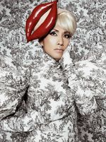 yozmit wears an outfit in the same 19th-century-pottery inspired print as the backdrop and a fascinator that looks like a huge mouth. yozmit has a concerned expression, with one hand under her chin and the other against her chest. She is an East Asian person with light-skin and platinum bangs peeking out from under the fascinator.