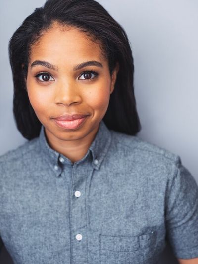 Carmen Lobue is a Non-Binary Afro-Philipinx shown from the upper chest up. Carmen is smiling wearing a blue-gray button up in front of a light blue background. They have brown eyes and light brown skin with straight dark brown hair going down their back.
