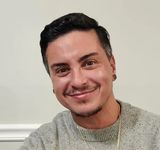 Elliott Feliciano smiling. Elliott is a Puerto Rican person with short dark hair with a little grey on the sides, light skin, and short facial hair. He wears small plug earrings, a gold necklace and a pullover.