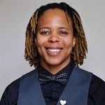 Evolve Benton is Black with brown skin wearing a navy blue button up and a slightly lighter blue vest with a navy blue and white polka dotted bow tie. They have ombre dark brown faded to light brown shoulder length dreads. They are wearing a heart pin on their vest that has the rainbow flag on it and dark brown earrings in front of a gray background.