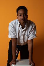 Hannah Patterson leans over a stool with a slight smile. Hannah is a Black person with dark skin and short hair. She wears a pinstripe button-up shirt and has a keychain hanging off her belt.