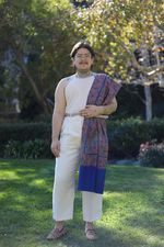 Kaith Karishma stands in a green space with a neutral expression. Kaith is a mixed Indian person with glasses, light skin, straight short hair, a mustache and tattoos on their neck, shoulders and left arm. They wear a white tank top, white pants, and a geometric print fabric drapped over their shoulder reminiscent of a sari.