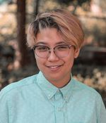 Kay Chan smiles in an outdoor setting. Kay is a Chinese and Métis person with glasses and ear-length blond hair with black roots. They wear a button-up with a subtle wave print.
