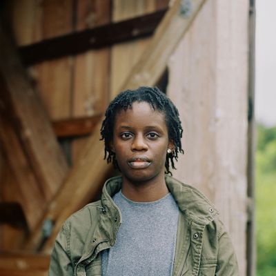 Kaydx with a neutral expression in front of a barn. Kaydx is a Black person with medium skin and short locks. Kaydx wears an army-green jacket over a t-shirt and has a stud earring.