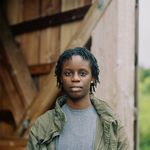 Kaydx with a neutral expression in front of a barn. Kaydx is a Black person with medium skin and short locks. Kaydx wears an army-green jacket over a t-shirt and has a stud earring.