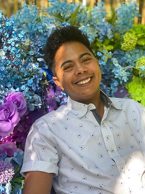 Ley Comas smiles in front of a giant bouquet of flowers. Ley is an Afro-Caribbean person with short curly hair, brown skin, and a nose ring. They wear a button-up shirt.
