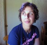 Lisset Barcellos gives side eyes while sitting in a chair. Lisset has wavy chin-length dark hair and light skin. They wear a t-shirt and a beaded necklace.