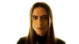 Sepand Mashiahof is a trans-femme Iranian-American in front of a bright white background. She has light skin and long straight brown hair. She is wearing a black turtleneck, a gold necklace and gold dangle earrings.