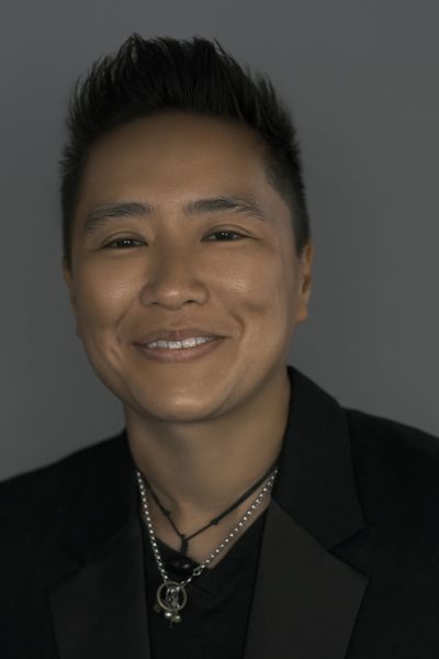 Sophia Shek is a nonbinary Chinese person with tan skin in front of a dark gray background. They are wearing a black suit jacket with a black t-shirt underneath. They have two necklaces, one black string and another silver chain with charms on it. They have short black hair faded on the side.