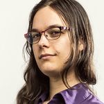 Tobi Hill-Meyer is an Indigenous Chicana trans woman with light tan skin in front of a white background. She is shown from the top of the shoulders up with a dark purple button up. She has long brown hair wearing small framed glasses.