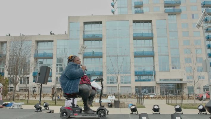 Mother LaTravious delivers a self-written poem at a Trans Day of Visibility event at Marsha P. Johnson Park in Brooklyn, New York. Mother LaTravious, A Black trans woman, holds a microphone and performs her poem on a small stage in a park on an overcast day. Her dark hair is in a protective hairstyle and she wears a fuzzy blue jacket and pleather black pants. She sits in a 4 wheel electric mobility scooter.