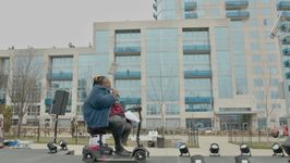 Mother LaTravious delivers a self-written poem at a Trans Day of Visibility event at Marsha P. Johnson Park in Brooklyn, New York. Mother LaTravious, A Black trans woman, holds a microphone and performs her poem on a small stage in a park on an overcast day. Her dark hair is in a protective hairstyle and she wears a fuzzy blue jacket and pleather black pants. She sits in a 4 wheel electric mobility scooter.