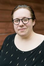 Jess Murwin is a nonbinary, mixed Indigenous settler with light skin. They have slicked back dark brown hair and are wearing black glasses. They are posing in front of a dark brown wooden background wearing a black t-shirt with small yellow lightning bolts across it.