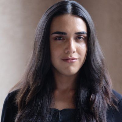 Ryan Rox is trans and non-binary with light skin. They have long straight brown hair and are posing in front of a tan background in a black blouse.