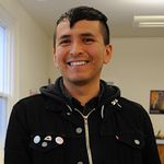 Demian DinéYazhi’ is a trans non-binary Indigenous person in front of a white background. They are wearing a black zip up jacket. He has short black hair with the sides buzzed.