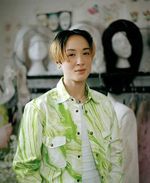 Sin Wai Kin is non binary and mixed race with light skin and posed in front of a blurred out closet space. They have ombre short hair with blonde ends and dark brown roots and a septum piercing. They are wearing a light green and white jacket with a white t-shirt underneath it with their hands in their pockets.