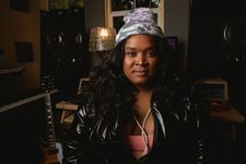 Leahann "Lafemmebear" Mitchell is a Black trans woman with brown skin posed in a music studio. She is wearing a gray and white beanie that says, You Stress Me Out. She has long curly black hair and is wearing a shiny zip up jacket with a pink tank top underneath and a long pearl necklace.