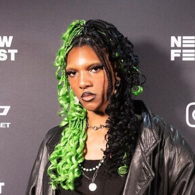 Clementine Narcisse is a Black trans woman with brown skin posed in front of a black banner. She has box braids in a half up half down style. Half of her hair is dyed lime green and the other half is black. She has three chain necklaces and is wearing a leather jacket with a black t-shirt underneath.