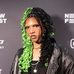 Clementine Narcisse is a Black trans woman with brown skin posed in front of a black banner. She has box braids in a half up half down style. Half of her hair is dyed lime green and the other half is black. She has three chain necklaces and is wearing a leather jacket with a black t-shirt underneath.