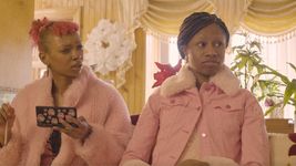 Two people sitting on a couch looking off to the right. They both have brown skin. The one on the left has pink hair with bangs and pulled back into a bun. They also have a pink furry coat on and a pink top on underneath. They are holding a black DSI gaming system covered in stickers in one hand and pink pencil in their other hand. The one on the right has brown box braids pulled into a low ponytail. They are wearing a pink denim jacket with a fur collar and fur cuffs. They have a nose piercing and a pink fur t shirt underneath and money sign earrings. Both people are looking at something off-camera to the right. The person on the left is bewildered by it, while the person on the right appears to be reacting with bemused acceptance.