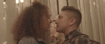 Two people stand really close together facing each other and the background is blurred out white street lamps. The person on the left has tan skin, shoulder length curly hair, wearing hoop earrings and a denim jacket. The person on the right has tan skin, short brown hair with the sides faded, wearing a flannel t shirt.