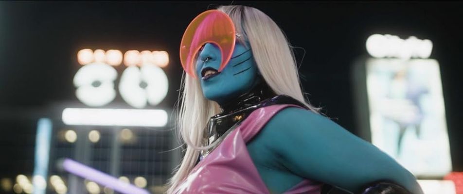 The person is posing in front of a blurred city building. They have blue skin, long straight blonde hair with bangs, and a pink latex top. They are wearing orange crescent shaped glasses.