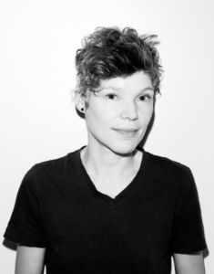 Classic, high quality black-and-white portrait of Nik Forrest against a bright white studio backdrop. Nik is a white trans person with short effortless wavy hair, small dark stud earrings and a fresh face. They wear a black V-neck t-shirt.