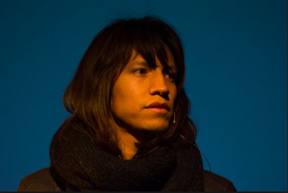 Close up of Beric listening attentively on a blue stage with a strong orange warm spotlight illuminating their face and hair. Beric is a medium light skinned Two Spirit person with a clean shaven face, dark brown facial features and medium long brown hair with a fringe. They are wearing a sleek professional headset microphone, a black long sleeve shirt, and chunky black knitted scarf.