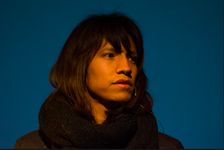 Close up of Beric listening attentively on a blue stage with a strong orange warm spotlight illuminating their face and hair. Beric is a medium light skinned Two Spirit person with a clean shaven face, dark brown facial features and medium long brown hair with a fringe. They are wearing a sleek professional headset microphone, a black long sleeve shirt, and chunky black knitted scarf.
