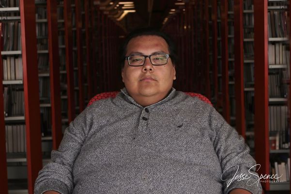 ODMK, an Indiginenous Two Spirit person, sits in a darkened library and stares directly into the camera with a neutral expression. They are wearing square glasses and a grey sweater.