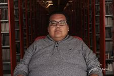 ODMK, an Indiginenous Two Spirit person, sits in a darkened library and stares directly into the camera with a neutral expression. They are wearing square glasses and a grey sweater.