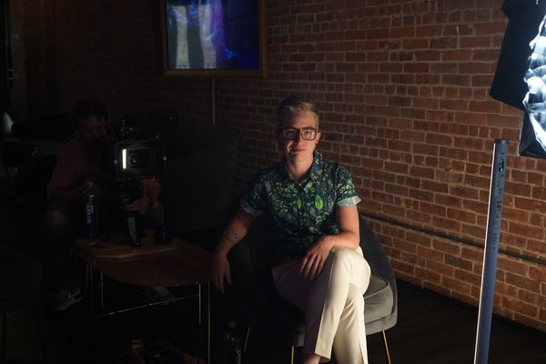 Behind-the-scenes photograph of Nico Opper on set. Nico has light skin, light salt-and-pepper hair in a short style, and hazel eyes under eyeglasses. They are seated in a chair in a casual pose for the camera, with filming equipment and crew in the background in shadows.