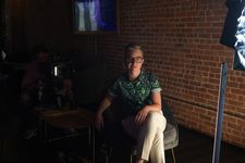 Behind-the-scenes photograph of Nico Opper on set. Nico has light skin, light salt-and-pepper hair in a short style, and hazel eyes under eyeglasses. They are seated in a chair in a casual pose for the camera, with filming equipment and crew in the background in shadows.