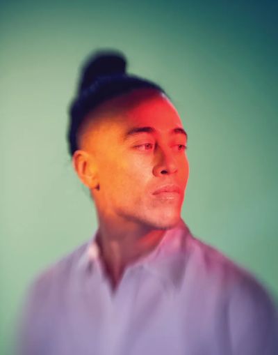 Wu Tsang has medium skin and dark hair tied up in a bun. She is wearing a light-colored shirt and looking off to the side with a contemplative expression. Creative and colourful aura lighting creates a soft, colorful gradient effect, blending shades of red and orange across the face and green in the background.