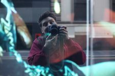 Mirror selfie using a professional camera of Asten Holmes-Elliott using the reflection on a city building. They have fair skin, thick, brown coiffed hair, and are wearing a sweater. The camera covers the lower half of their face, but an excited smile is visible through their wide eyes and raised eyebrows. An abstract visual of some kind appears to be overlaid on the image.