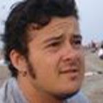 Cropped image showing Mikajlo Ranković from the shoulders up. They are outdoors, on what appears to be a beach. He has tanned skin, dark brown hair and light facial hair.