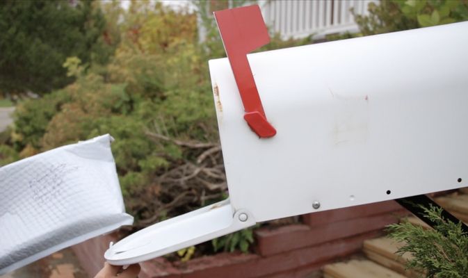 A hand opening an old-school white tin mailbox with a red flag. In the other hand, which is out of frame, is a parcel with handwriting on it. Green bushes and a white railing are in the background.