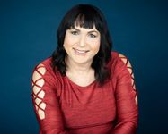 Headshot of a light-skinned, brown-eyed trans woman with black hair below her shoulders and bangs. She smiles kindly at the camera. She is wearing a red top with laces on the shoulders and upper arms.