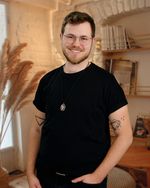 Image of Markus Harwood-Jones, a white, bearded, trans masculine individual wearing glasses, in a black shirt, smiling in front of a bookshelf.