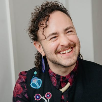 Image of Shane Forrest, an Anishinaabe-Métis Two-Spirit person with light skin, curly brown hair, a mustache, and some stubble along their chin. They wear a dark vest covered in colorful patches, earrings, and smile widely just past the camera.