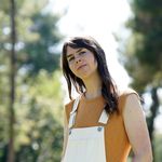 Low-camera angle of Sarah Wheldon on a sunny day in a park. Sarah has light skin and brown hair falling past her shoulders. She wears white overalls and an orange t-shirt.