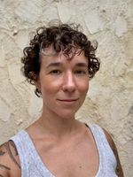 Selfie taken by Chelsea Moore from the shoulders and above in front of a textured outdoor wall. Chelsea has fair, tanned skin, and a dark curly bob with a blonde accent piece by her face. They are wearing a grey tank top and she has a thin silver nose ring.