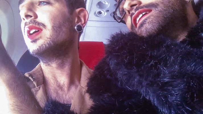 Landscape iPhone footage of two Palestinian drag queens, Elias Wakeem and Oz Marinov, seated in an airplane looking out of the window, illuminated by the daylight. Their faces are not fully in the image, but their mouths are slightly open in an awed expression. They are both in drag, wearing fur, red lipstick and earrings.