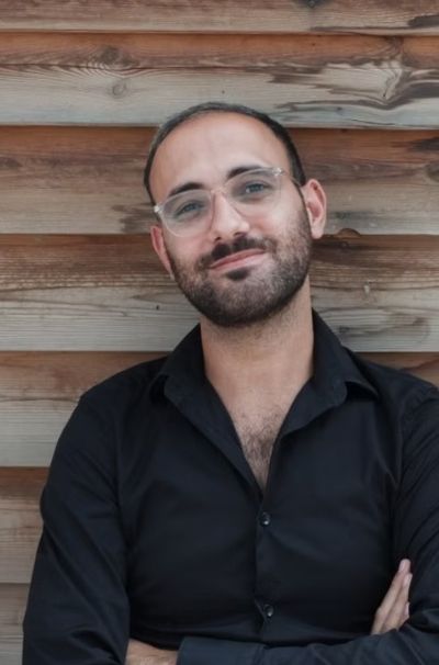 Classy portrait of Elias Wakeem, an Arab genderqueer person, standing in front of a wooden wall which appears to be outdoors. Elias is wearing a black button up shirt with the top button opened and clear glasses. Elias has dark facial hair and a short haircut.