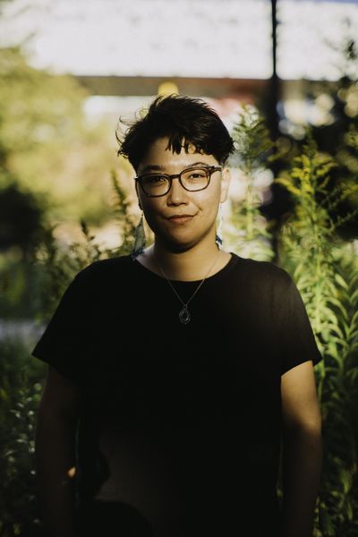 Kay looks directly at the camera with a confident yet sly smile. They have light skin, short dark hair, and dark brown eyes. They are wearing dark grey glasses, a black shirt, and a necklace. Behind htem are some blurry young trees.