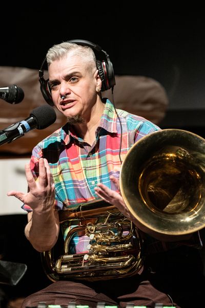 Lucas Crawford speaks expressively into a microphone while holding a tuba. He was fair skin, short, styled gray hair, a dark goatee, a nose ring, and wears a rainbow-coloured plaid button-up shirt.